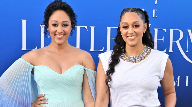 Tamera and Tia Mowry posing and smiling in front of a blue Little Mermaid background.