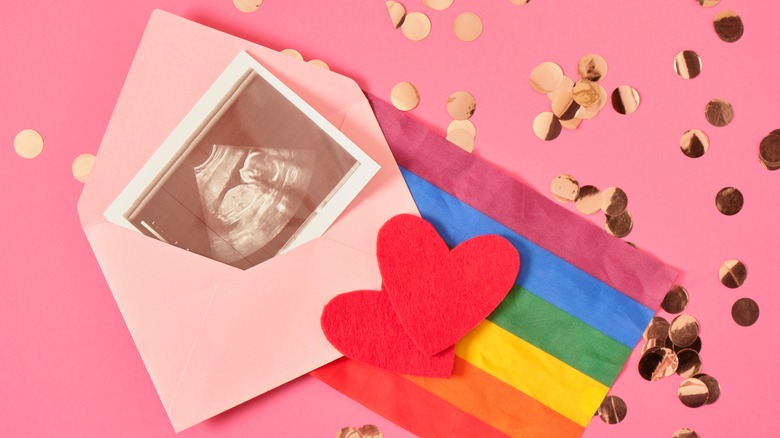 Open envelope revealing sonogram with rainbow tissue paper, red hearts, and gold confetti
