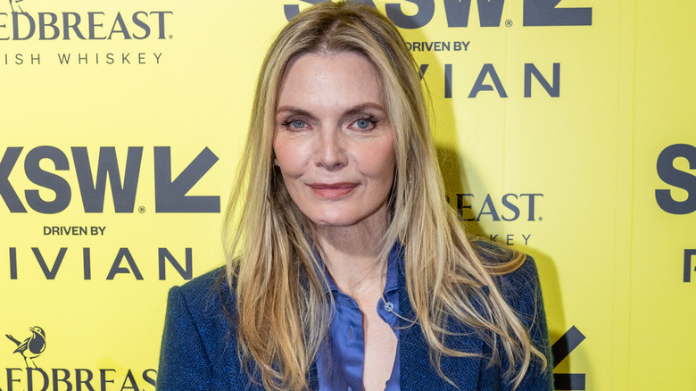 Michelle Pfeiffer smirking while posing at an event against a yellow and black background