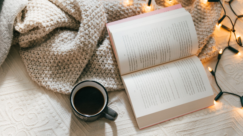 book open on a bed with lights and a blanket next to a mug