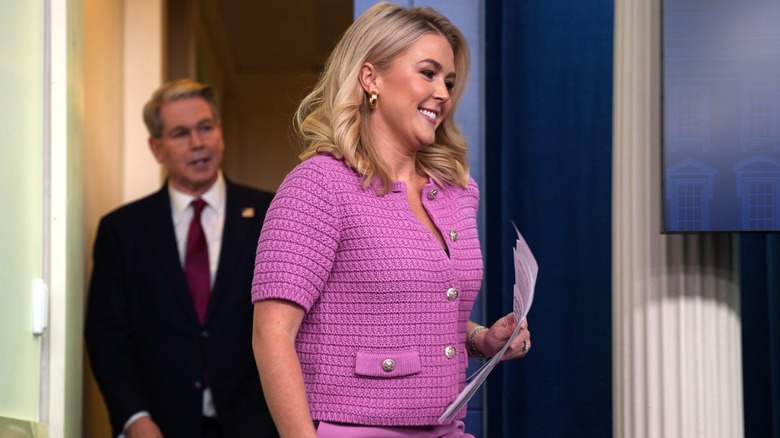 Karoline Leavitt in the White House press room wearing a knitted purple jacket and matching pants, with a man stood behind her