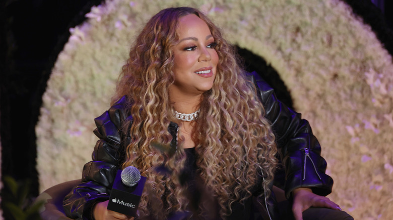 Mariah Carey holding a microphone onstage in a leather jacket with curly hair