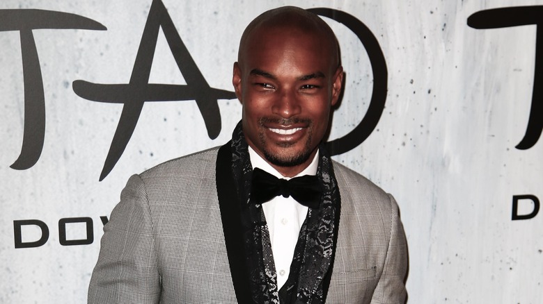 Tyson Beckford smiles in a gray suit with a black scarf and matching tie.