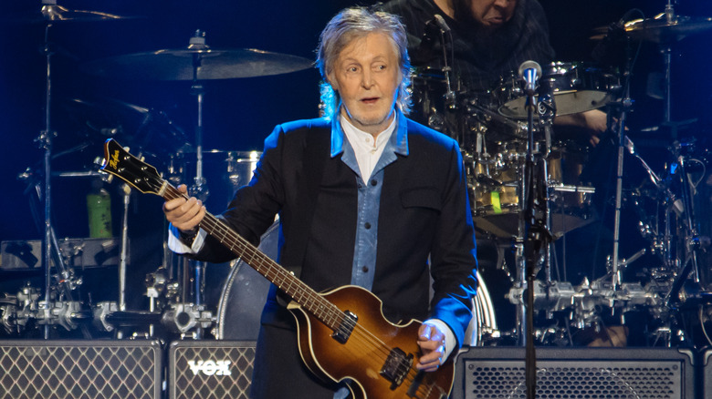 Paul McCartney playing bass guitar onstage in a denim shirt and black jacket
