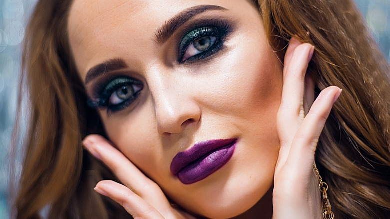 Woman with crimped hair wearing dramatic eye makeup and lipstick touching her face and  tilting her head to the side.