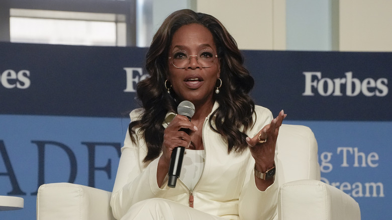 Oprah Winfrey wearing white and sitting on a couch on a stage holding a microphone