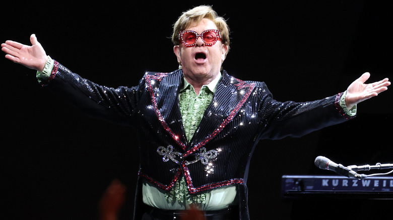 Elton John onstage with his arms extended in a glitzy jacket and sunglasses