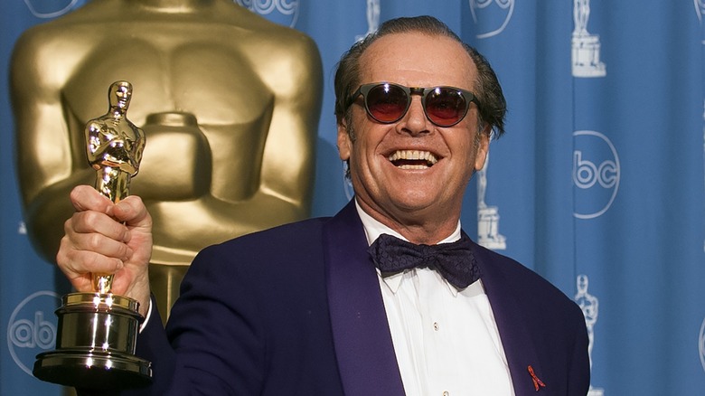 Jack Nicholson smiling while holding up his Oscar in a purple jacket, matching bow-tie, and sunglasses