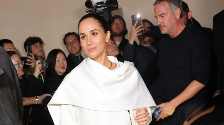Meghan Markle walking in a white wrap surrounded by people photographing her