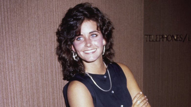 Young Courteney Cox looking over her shoulder to the left in front of a brown background.