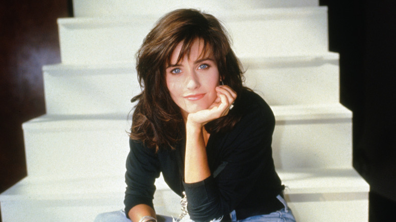 Courteney Cox props her head on her hand, sitting on a set of white stairs.