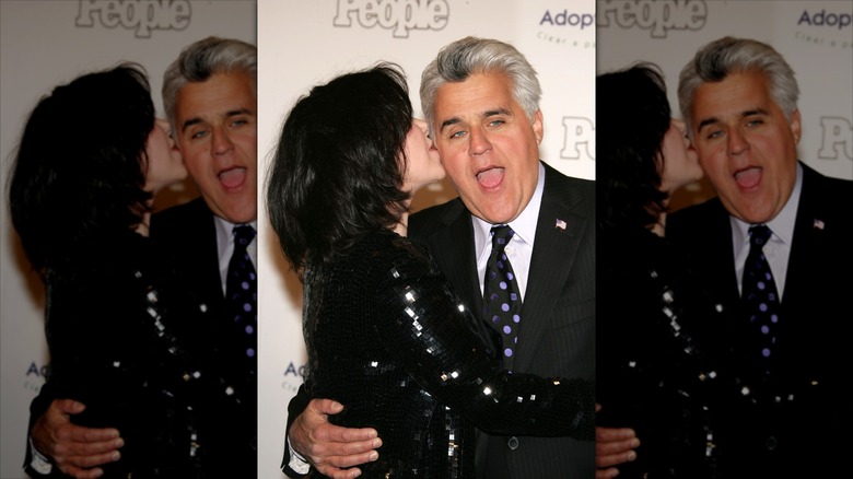 Mavis Leno with her mouth to Jay Leno's ear as he makes a shocked expression