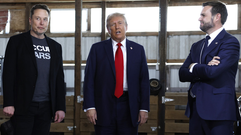 Elon Musk, Donald Trump, and JD Vance standing together in front of a window, with Trump talking and Vance looking at Musk