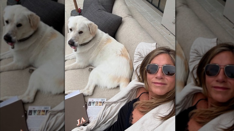 Jennifer Aniston wearing sunglasses, a white robe and black top while smiling next to her dog.