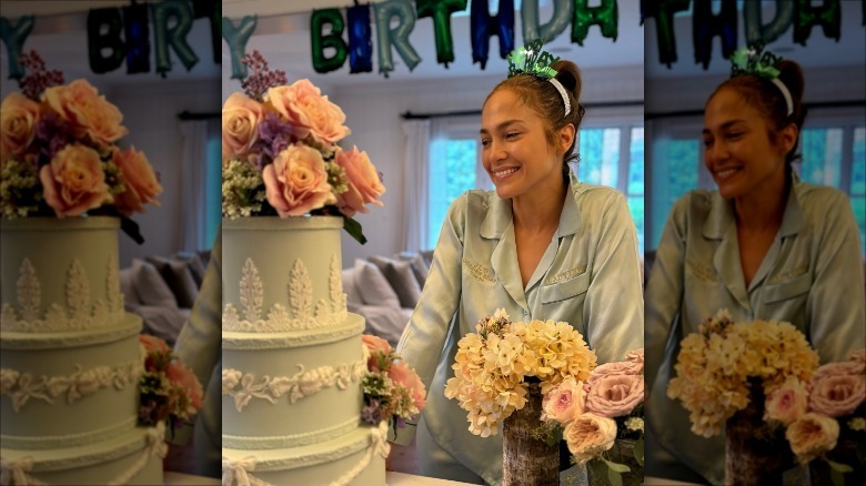 Jennifer Lopez smiling at a birthday cake while wearing birthday pjs and a "Happy Birthday" crown in front of "Happy Birthday" balloons