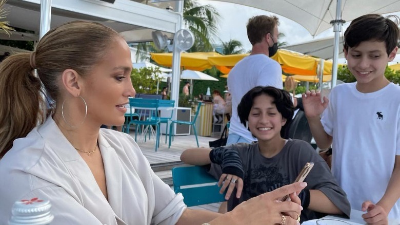 J.Lo looking at her phone while Emme and Max Muñiz smile next to her