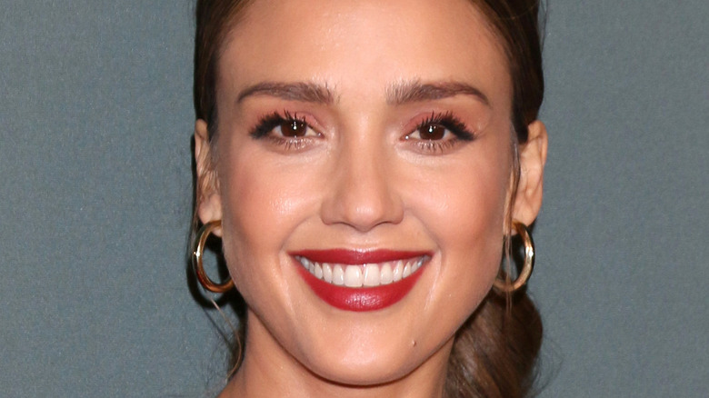 Jessica Alba smiles with red lipstick and hoop earrings.