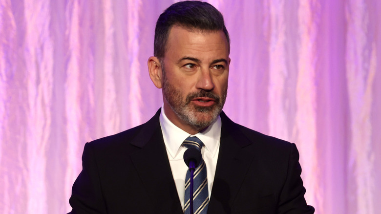 Jimmy Kimmel delivering a speech in a suit in front of a purple backdrop