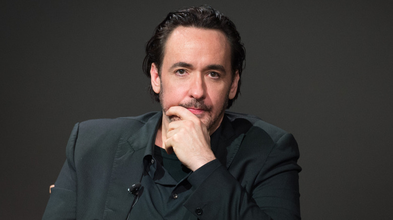 John Cusack sitting with his hand on his chin