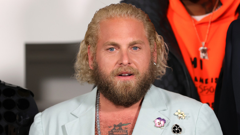 Jonah Hill, exposing his tattoo-covered chest, beneath a pale blue blazer with flower pins on the left shoulder