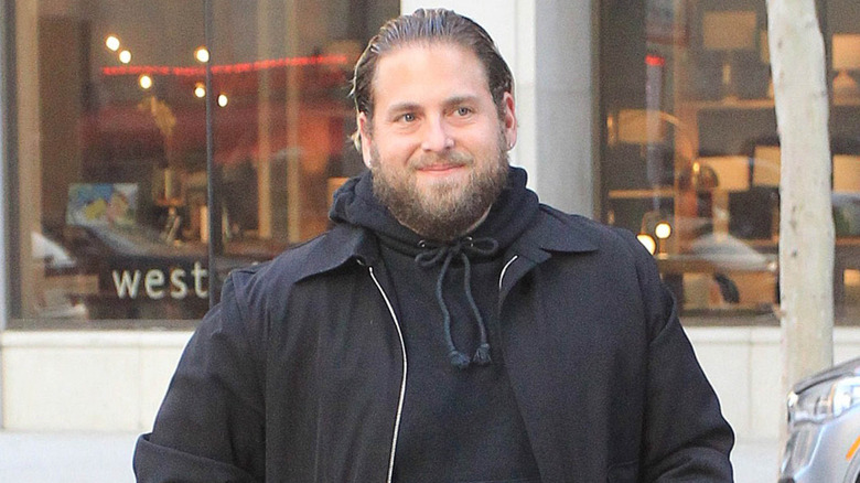 Jonah Hill, smiling on the street, in black jacket over hoodie