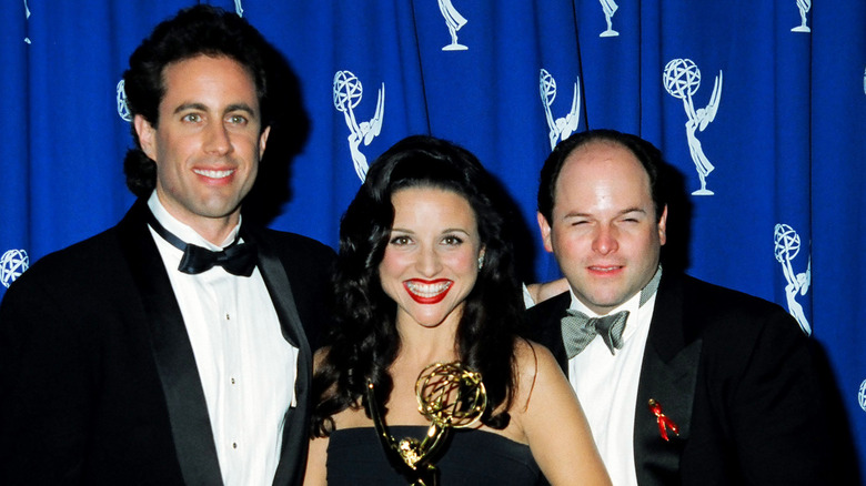 Julia Louis-Dreyfus smiling while holding an Emmy award, flanked by Jerry Seinfeld and Jason Alexander in tuxedos