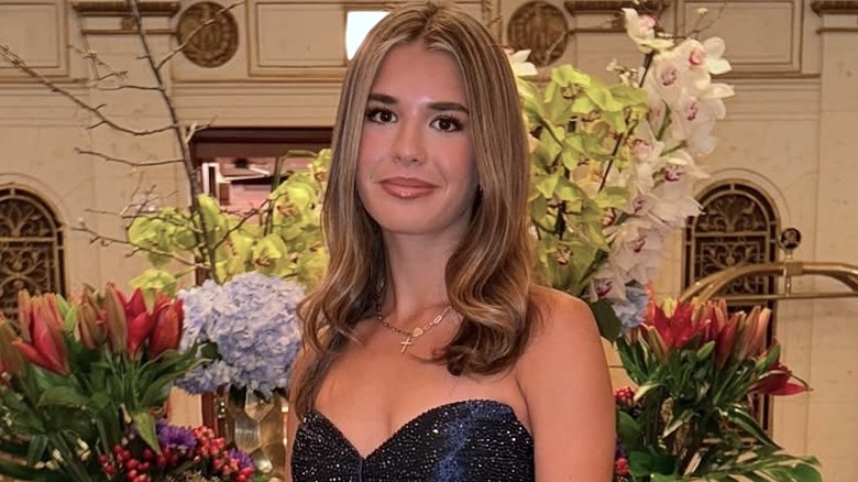 Kai Trump standing in front of flowers, wearing a strapless sparkly blue gown, lightly smiling