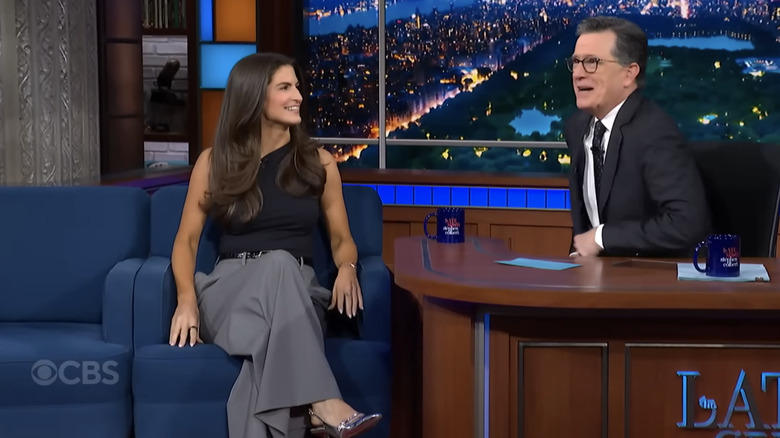 Kaitlan Collins, sitting on a sofa chair, next to Stephen Colbert, behind a desk