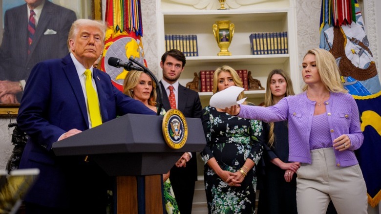 Karoline Leavitt hands Donald Trump a document as he speaks in the Oval Office (2025)