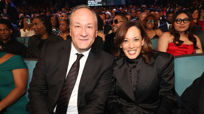 Doug Emhoff and Kamala Harris smile sitting in the audience of an event