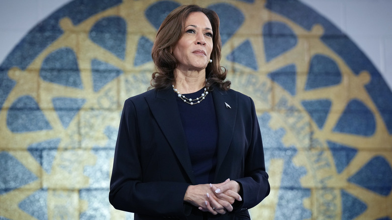 Kamala Harris standing with her hands folded in front of a yellow and blue mural (2024).