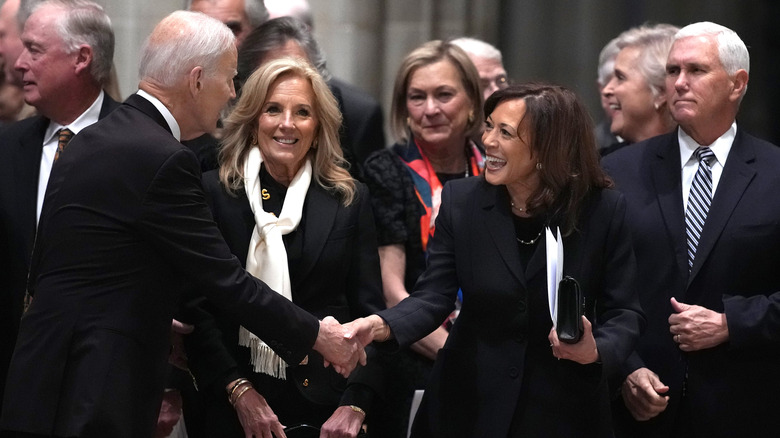 Kamala Harris smiling and shaking Joe Biden's hand with Jill Biden in the middle
