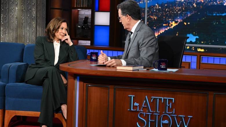 Kamala Harris on the set of "The Late Show with Stephen Colbert," leaning on Colbert's desk and listening to him as he talks.