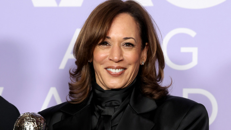 Kamala Harris dressed in black, smiling in front of a purple background.