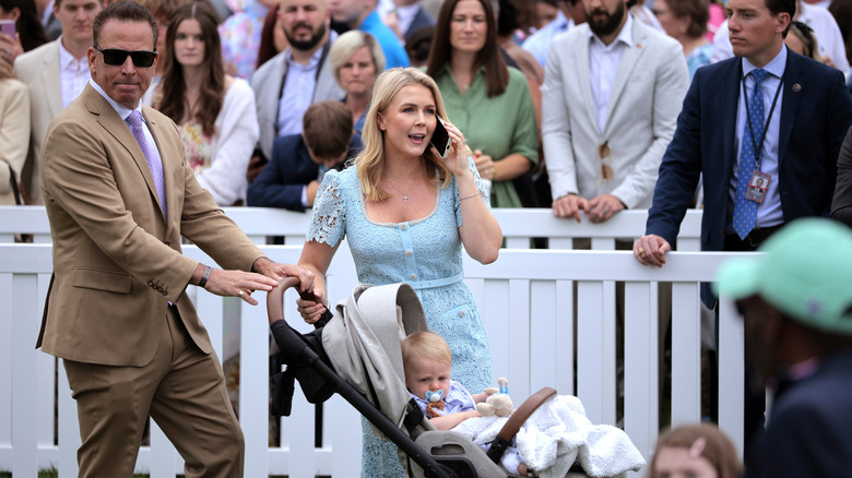 Karoline Leavitt speaking on the phone in a light blue dress while Nicholas Riccio pushes their toddler in a stroller
