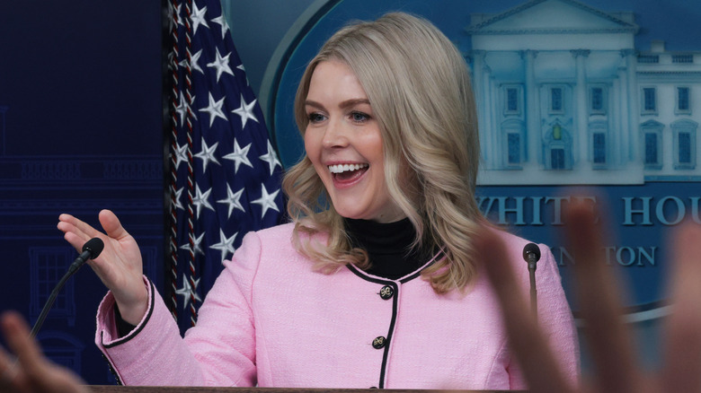 Karoline Leavitt gesturing at the podium in a pink collarless jacket
