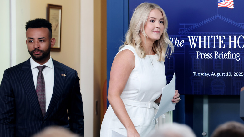 Karoline Leavitt smiling as she walks to the White House podium in a sleeveless white dress