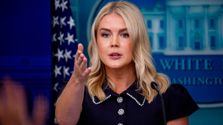 Karoline Leavitt addressing the press in a black dress while gesturing with her right hand