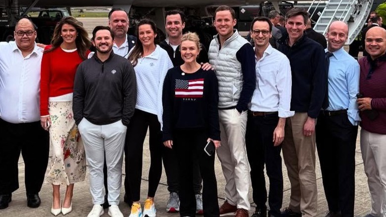 A group of people pose together in front of Air Force One, Karoline Leavitt is in the middle wearing an American flag sweater that reads "Trump" (2025)