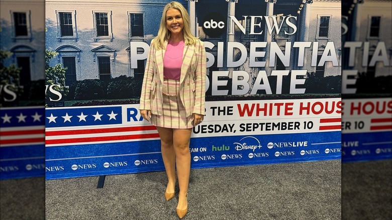 Karoline Leavitt standing in front of presidential debate poster wearing a matching plaid mini skirt and jacket