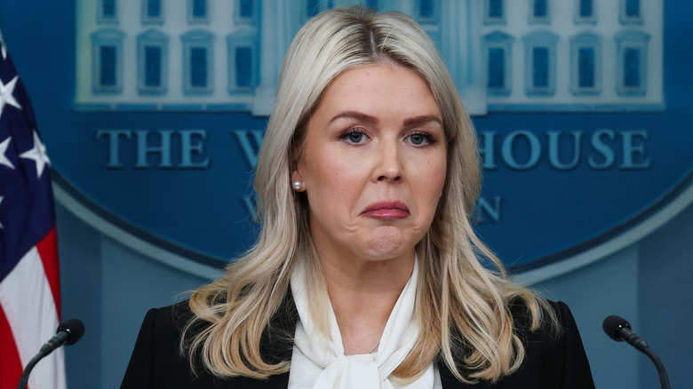 Karoline Leavitt making a sad face at the White House press podium in a black blazer with a white blouse underneath