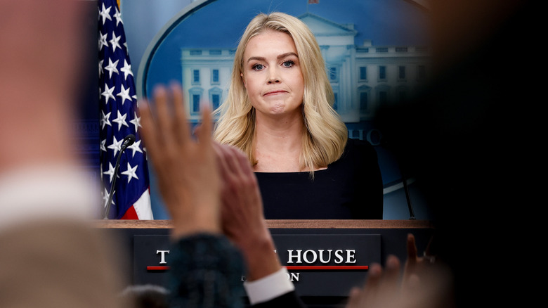 Karoline Leavitt frowning before a white house press room filled with raised hands.