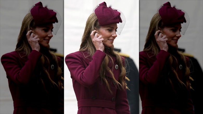 Kate Middleton puts her hand up to her face in a burgundy outfit with a matching hat