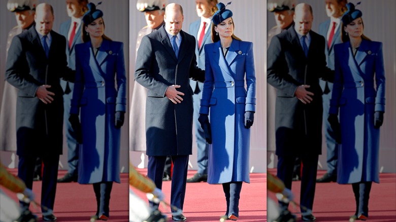 Prince William in a long blue coat with his arm on Kate Middleton's back at an outdoor event