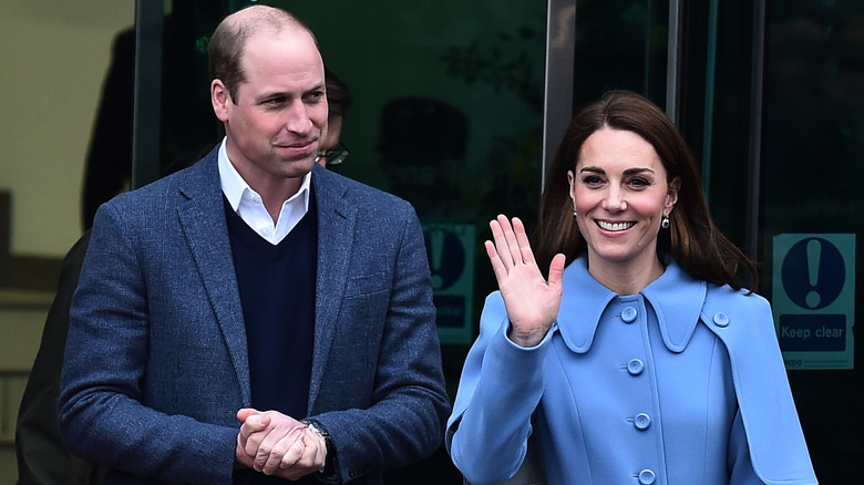 Prince William in blue with his hands clasped and Kate Middleton in blue waving