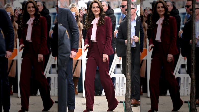 Kate Middleton smiling in a crowd wearing a burgundy pantsuit with a pink bow shirt underneath