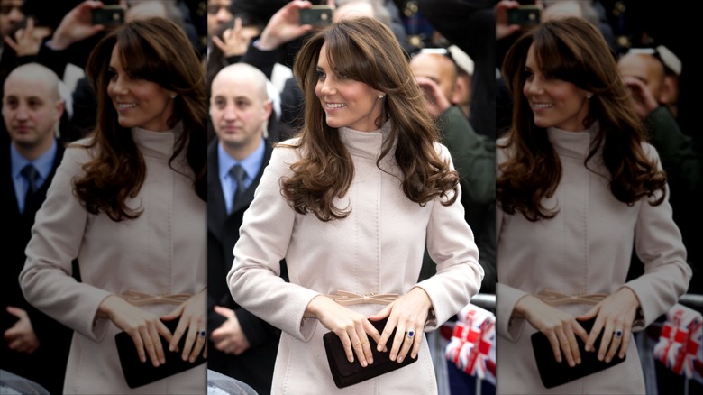Kate Middleton clutching a black purse, wearing a pink coat while smiling over her shoulder.