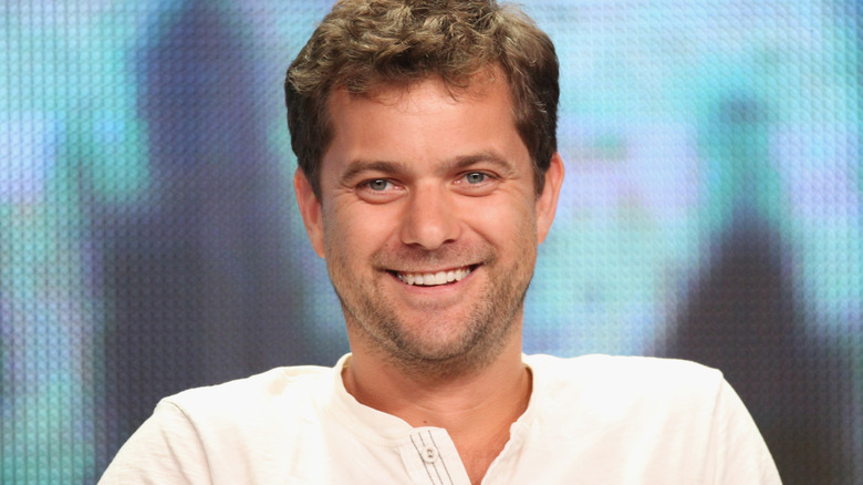 Joshua Jackson smiles at a camera while sitting against a blue backdrop
