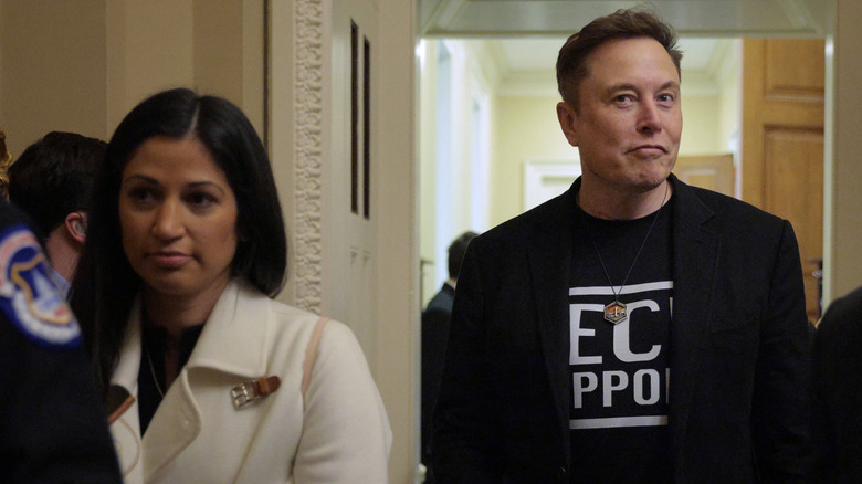 Elon Musk smirking while Katie Miller stands next to him speaking to someone