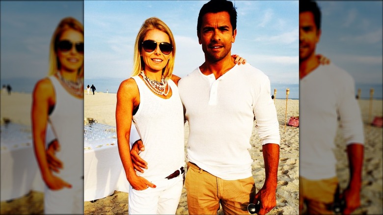 Kelly Ripa wearing a white tank top and pants and posing with arm around Mark Consuelos, who is wearing a white shirt and khakis